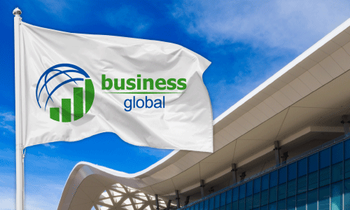 White fabric flag with blue sky and tan trade show venue roof in the background. Flag has a circular logo of a blue half sphere and green bar graph. Text next to the logo says Business in green lettering, and it says Global in blue lettering underneath.