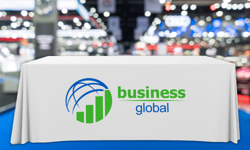 White table cover with circular logo of half a blue  sphere and a green bar graph below. Text beneath reads Business in green lettering, with Global in blue lettering underneath. Table is featured on blue carpeting with a blurred out trade show floor behind it.