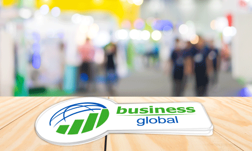 Die-cut vinyl decal with a circular logo of half a blue sphere and a green bar graph below. Text next to the logo reads Business in green lettering, with Global in blue lettering underneath. Decal is sitting on a white pine table with a blurred out trade show scene in the background.